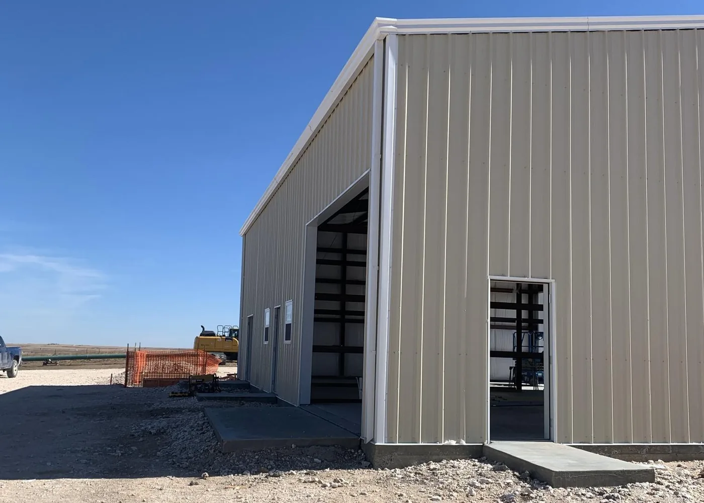 Tan steel workshop building under a clear blue sky, with a large open bay door and a walk-through door on a fresh concrete slab.