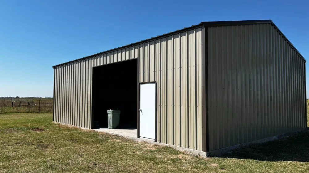 Dark green steel storage building with an open bay and white walk door in a grass field