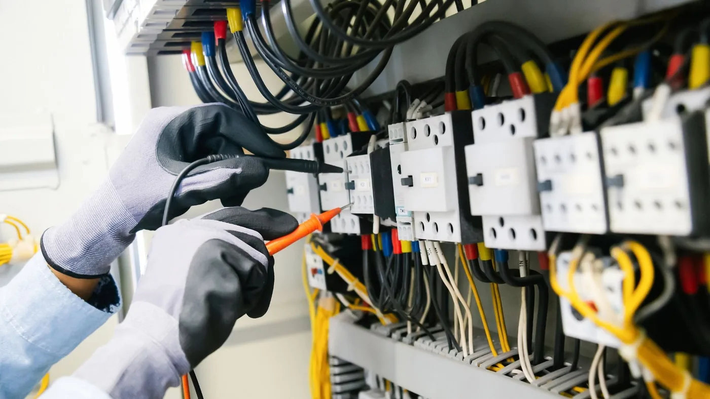 Gloved hands testing wiring and breakers inside an electrical panel with a multimeter
