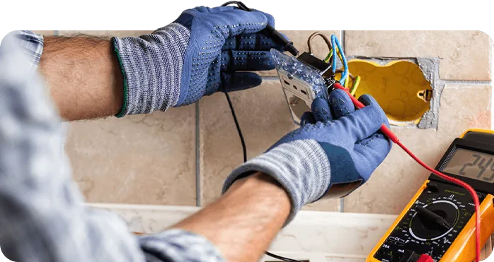 Electrician in work gloves wiring an outlet in a wall box beside a multimeter