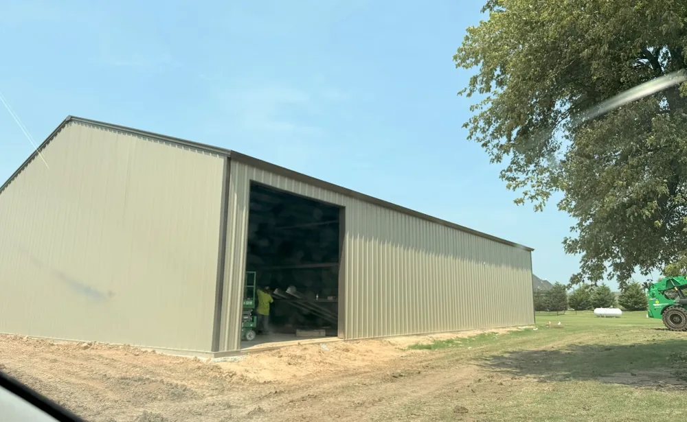 Tan metal shop building beside a farm drive with equipment parked under the trees
